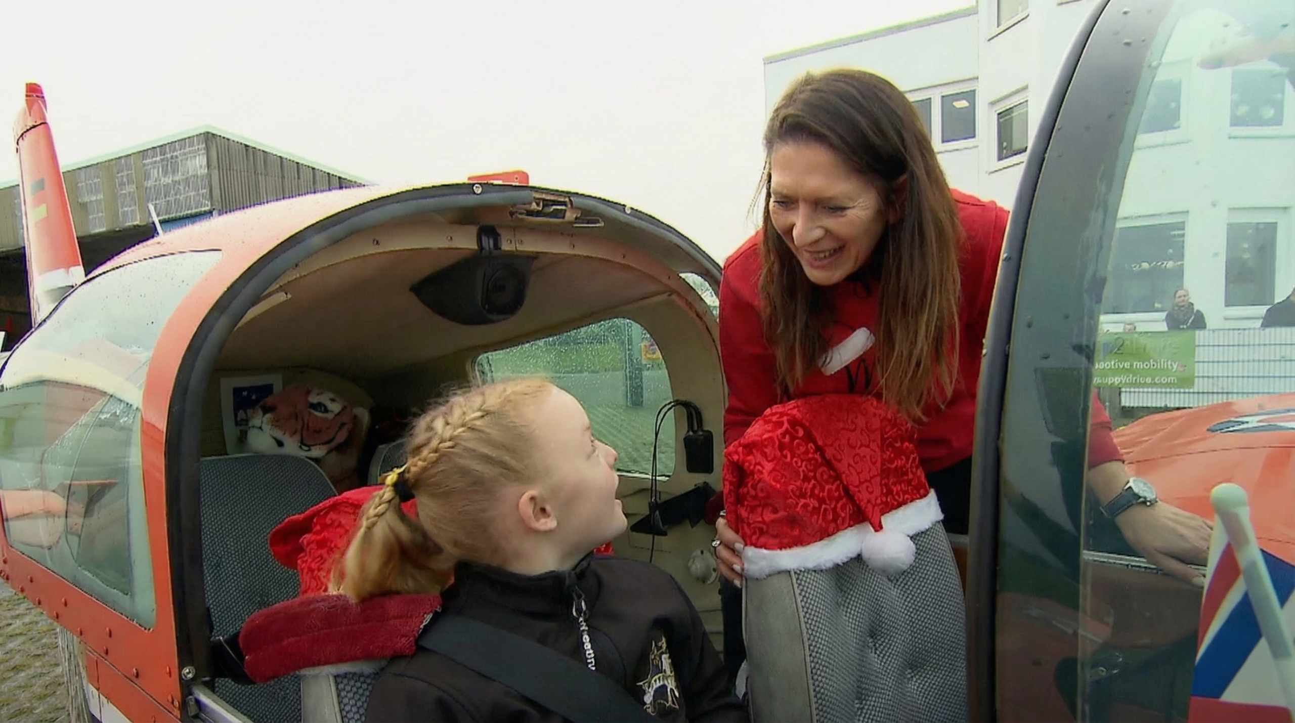 Pilotin Kathrin Kaiser lächelt ein Kind an, das im Cockpit eines roten Sportflugzeugs sitzt. Auf dem Sitz liegt eine Nikolausmütze. Im Hintergrund sind Gebäude des Flugplatzes Bonn-Hangelar zu sehen.